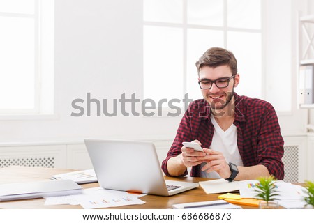 Young happy businessman using mobile by the laptop in modern white office interior.