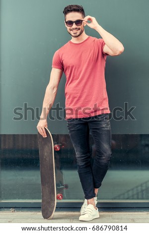 Stylish guy in sun glasses is holding a skateboard, looking at camera and smiling while standing outdoors, full length