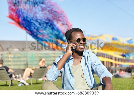 Outdoor picture of young dark-skinned male wearing trendy shades, hat and shirt sitting at green lawn phoning his friend having conversation with him smiling pleasantly while looking into distance