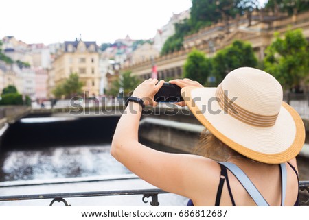 Woman taking pictures with smartphone. Stylish summer traveler woman in hat with camera outdoors in european city, old town Karlovy Vary in the background, Czech Republic, Europe