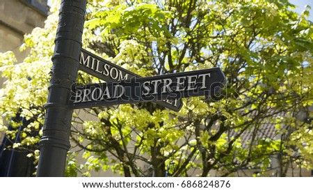 Road sign for Broad Street and Milsom Street in the city of Bath Spa, Somerset, England, with leafy green trees in the background, and bright sunlight