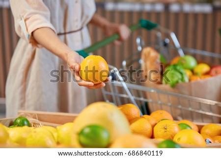 The hand of unidentified woman holding orange standing next to  vegetable stand and with cart full of useful organic super food
