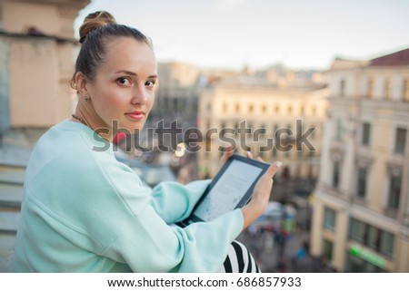 Attractive and stylish girl sits on the roof of the house in the old town and reads the e-book.