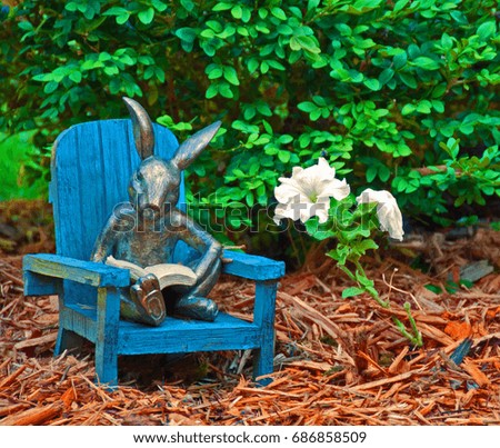 A photo of a garden ornament sitting in a mulched flowerbed - a rabbit reading a book in an Adirondack chair 