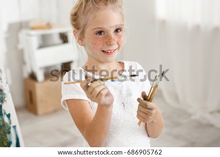 Close-up portrait of blonde cute European little girl with paint on her freckled face and hair bun smiling with all her teeth holding a bunch of brushes in her hands. Cheerful girl messed up her white