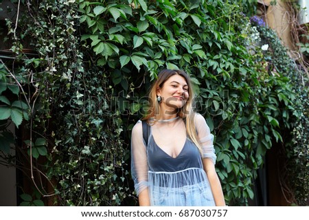 Outdoor lifestyle picture of happy relaxed young Caucasian blonde female student in transparent dress and earrings having cheerful pleased look, spending free time after college in urban park