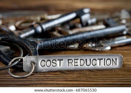 Photo of key bunch on wooden board and tag with letters imprinted on clean metal surface; concept of COST REDUCTION