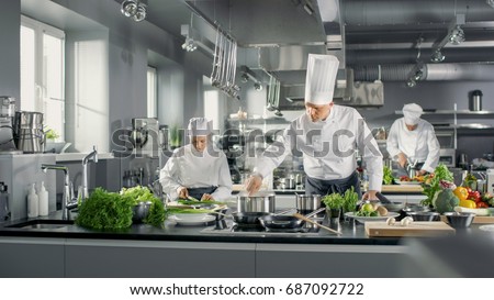 Famous Chef Works in a Big Restaurant Kitchen with His Help. Kitchen is Full of Food, Vegetables and Boiling Dishes. Royalty-Free Stock Photo #687092722