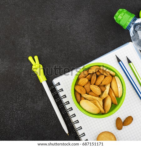 School lunch in a green round container with cookies and almonds on a dark background with handles and notebooks, pencils. Top View.