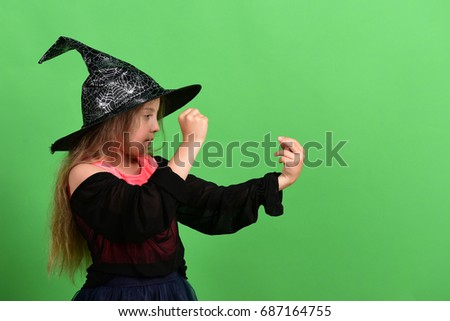 Kid in witch hat, dress and concentrated face. Girl tries to put spell, isolated on green background. Child in witch costume black hat. Halloween and celebration concept