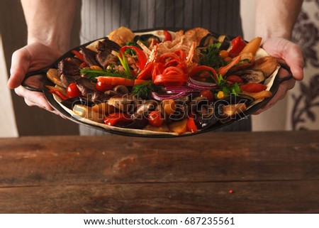 Presentation dish with stewed meat, different kind of vegetables, field mushrooms, laid on lavash and big copper plate, and decorated with sliced onion and pepper. Meal in hands