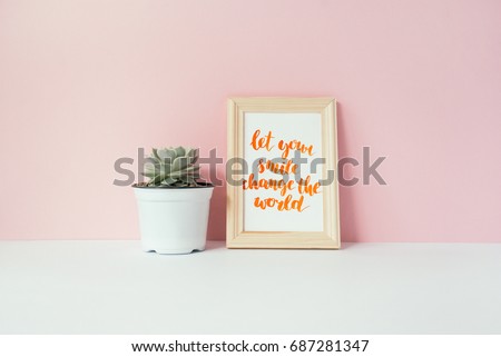 Succulent in white pot and card with inspirational quote "let your smile change the world" in a wooden photo frame in front of pale pink pastel background