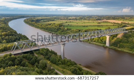 A bridge crossing the Kiel Canal (German: Nord-Ostsee-Kanal) in Schleswig Holstein in Germany with lush green fields and wind farms in the background is captured aerial from a drone at sunset. Royalty-Free Stock Photo #687317257