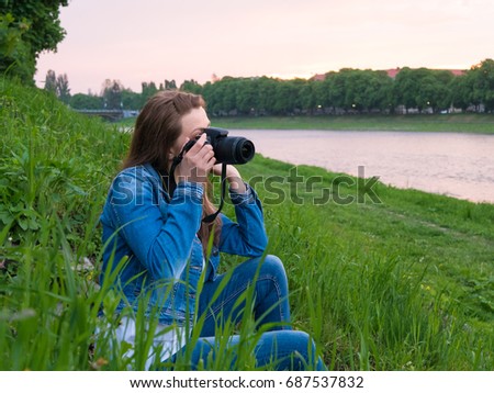 Beautiful girl tourist in a cotton jacket taking photos with a professional camera on the banks of the river in windy weather.