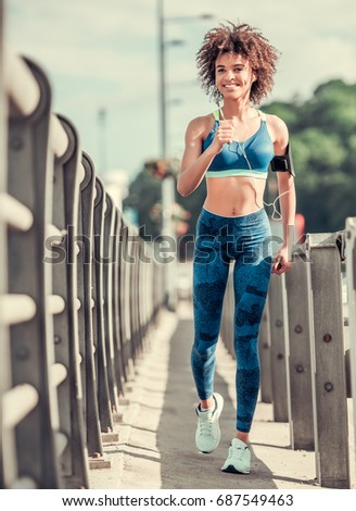 Beautiful Afro American girl in sportswear and earphones is looking at camera and smiling while running