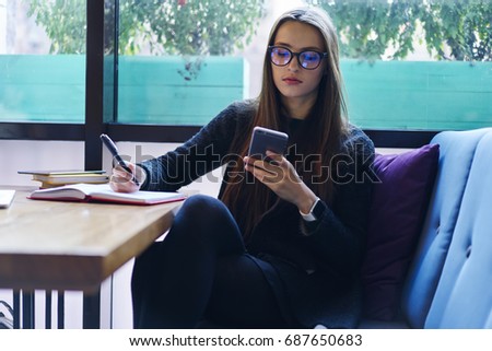 Pensive brunette young woman in eyewear noting information in diary while reading news via smartphone.Beautiful female student booking tickets and making payment on web page sitting in coffee shop