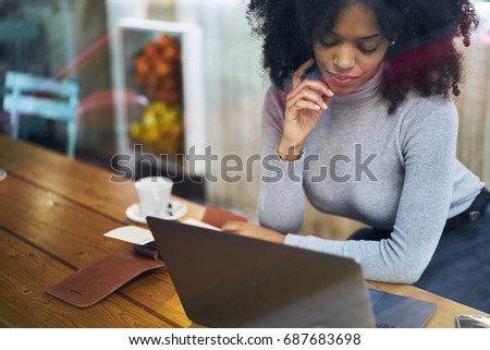 Pondering young afro american businesswoman dressed in trendy outfit watching training webinar on laptop connected to 4G.Thoughtful good looking female blogger sitting in coffee shop with gadget
