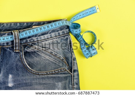 Healthy lifestyle and dieting concept. Upper part of denim trousers isolated on yellow background. Close up of jeans with measure tape around waist.  Blue jeans with blue measure tape instead of belt.
