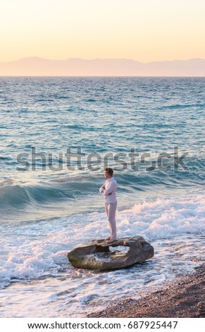 Happy carefree man in the white shirt and linen trousers stands barefoot on the rock close to the stormy sea. Male model experiencing emotion of freedom in the evening during the vacation. 