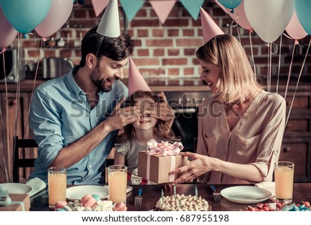 Happy family is sitting at the table in decorated kitchen during birthday celebration, dad is covering daughter's eyes while mom is holding a present
