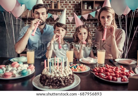 Happy family is blowing in party horns at camera while sitting at the table in decorated kitchen during birthday celebration