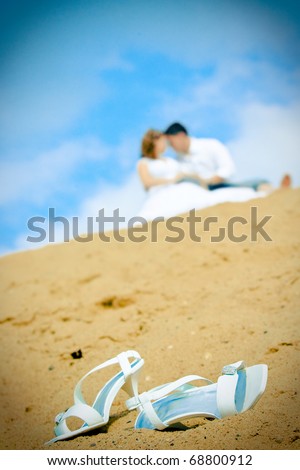 A close up of a bride's wedding shoes on the sand at the seaside with the couple in the background, focus on the shoes in front.