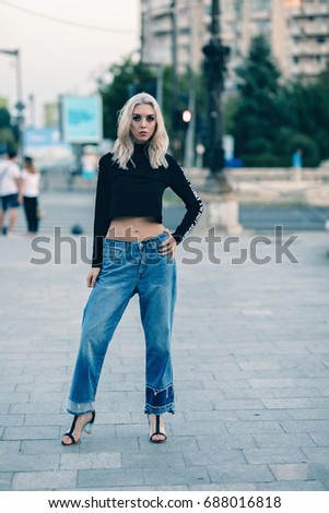 Posh girl posing for photo in twilight on tiled floor. Dressed in black full sleeve crop top and blue jeans with high heels. Couple in background and road is empty. Standing front of junction.