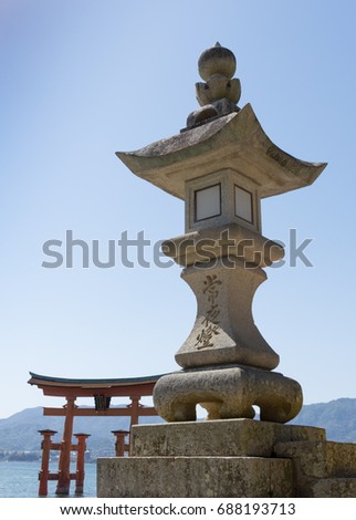 Close up of an engraved concrete shrine with the Torii Gate of Miyajima in the background. A small portion of the Bay of Hiroshima and a forested hillside are seen in the background. 