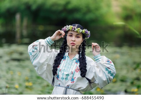 Cute brunette girl  with two pigtails hair braids wearing national Ukrainian costume and crown,  walking near the lake with bulrushes. 