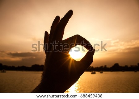 silhouette,The hand symbol signifies an ok, with the tip of the thumb and fingertips touching each other, with the background color being the orange sky and the lake.