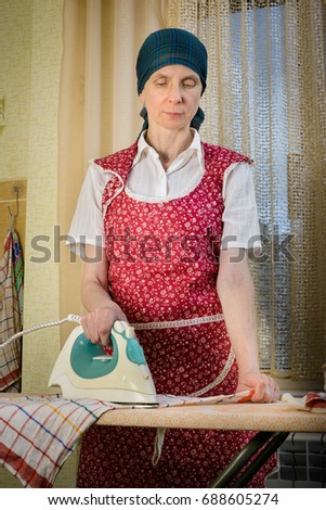 An adult woman, a housewife or a maid, with a green scarf on the head and wearing a red apron, is standing behind the ironing board. She irons some tea towels in the kitchen