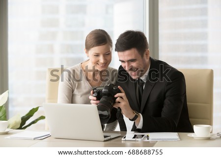Cheerful laughing office people holding professional camera, happy businessman showing pictures from photo shooting to businesswoman, colleagues having fun while watching shots of corporate party