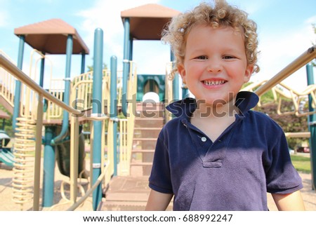 A little boy is smiling at the playground. The young boy has blonde curly hair and looks happy to be at the park. 