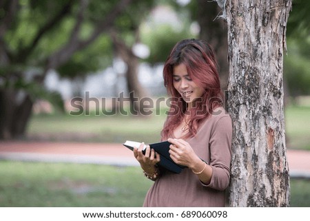 Portrait of happy charming young asian woman reading a book and listening music outdoors,sitting on grass looking something in the park
