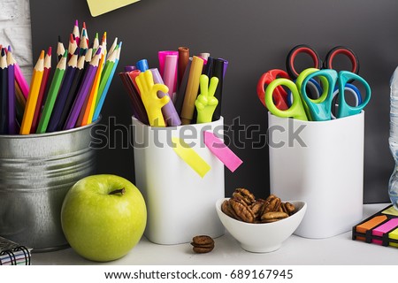 School background: a white brick wall with accessories, pencils, tetrads, scissors, an apple, pecans, a board for a substitute placed in front of her on the table. Selective focus