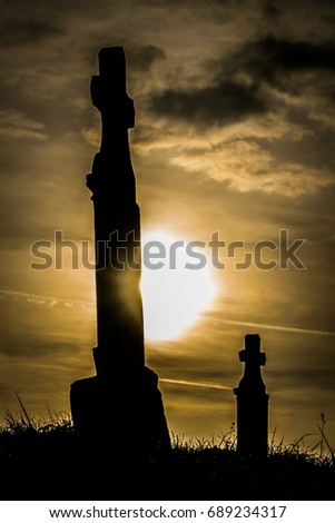 Graveyard at sunset dark clouds golden light
Cemetery at sunset with the silhouette of two tombstones with crosses

