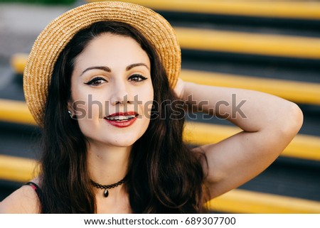 Pretty woman with dark hair, shining eyes and red painted lips looking confidently and with smile at camera wearing straw hat and necklace on neck. Positive female model having summer vacations