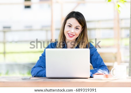 A beautiful young girl smiling and looking towards the camera while using a computer laptop outdoors.