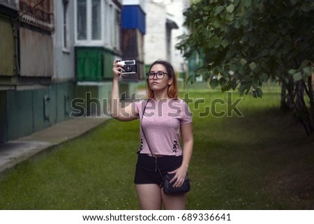 A young fashionable girl, a student, wearing glasses, wearing a pink T-shirt, black shorts, with a wallet walking in the street, taking pictures of nature on an old film camera in a green courtyard.