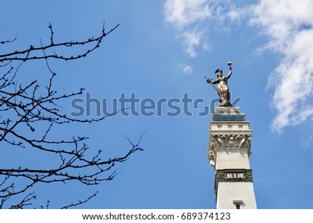 Lady Victory Statue in Monument Circle downtown Indianapolis with blue sky and bare tree branches in foreground.