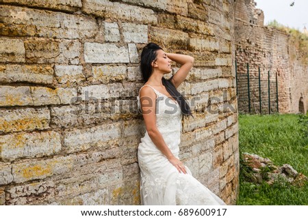 Young brunette woman in a white dress posing in front of an ancient fortress wall with one hand in her hair 