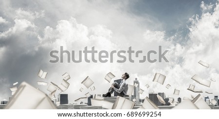 Businessman looking away while sitting on pile of documents among flying books with cloudly sky on background. Mixed media.