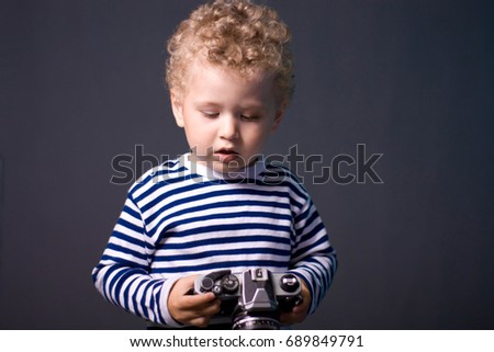 A small curly-haired boy with a camera on a gray background - a studio photo. The kid learns to take pictures.