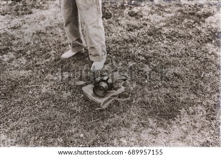Old sepia photo of man standing next to leather bag with gloves and camera lying in field. High angle view.