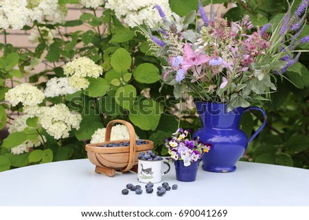 Flowers in vintage enamel jug, blue berries in vintage mini basket, enamel small mug, violet pansies in mini bucket on table, garden scene on hydrangea bush background, daylight, outdoors,