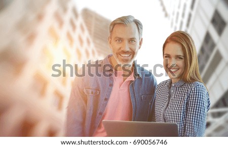 Smiling businessman and businesswoman are holding a laptop  against low angle view of office towers