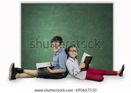 Picture of two elementary students looks happy while sitting near a blank chalkboard and holding a book in the studio