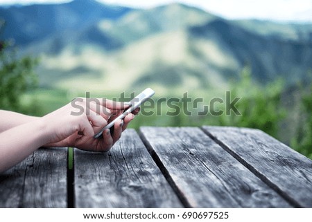 Woman's hands using mobile phone sitting at the old wooden table among the mountains. Green mountains on the background, outdoors, 