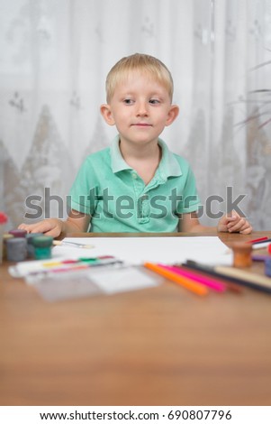 A little blond boy with blue eyes draws colored pencils on paper, a blank sheet on the table