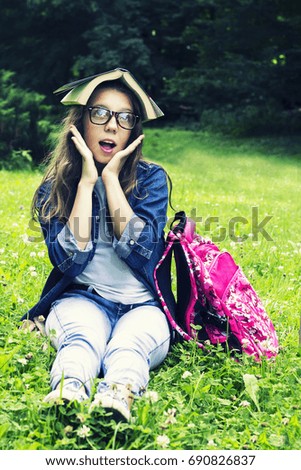 Beautiful blonde schoolgirl girl in jeans shirt reading a book on grass with a backpack in the park